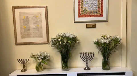 Three vases of white and green flowers and two Hanukkiah candlesticks sitting on a white shelf below framed excepts from the Jewish Tanakh.
