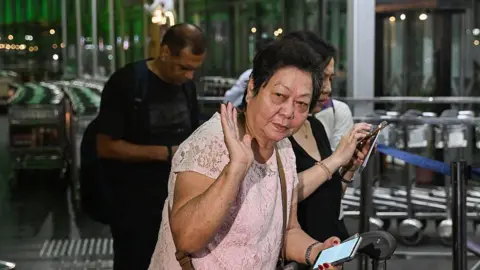 Getty Images A middle-aged woman with short black hair in a pink shirt waves as she looks to the side, at the entrance of an airport