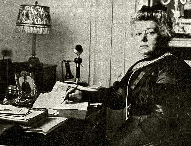 A black-and-white photograph shows a woman seated at a desk with papers and a lamp, looking directly at the camera while holding a pen.
