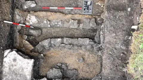 ODAIT A small square burial site is a mixture of brown mud and grey stone. There are two red and white thin sticks lying horizontally and parallel to each other. 