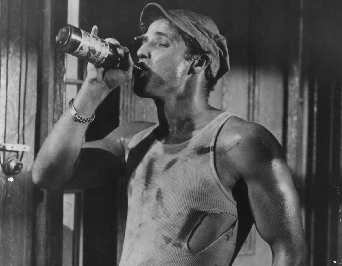 A black and white image of a man drinking from a bottle while wearing a hat, a torn and dirty sleeveless white-ribbed top and jeans. 
