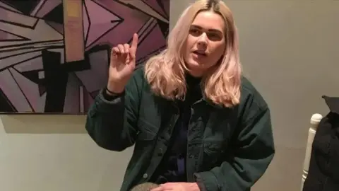 PA/Cardiff School of Journalism Bethan Roper sits in front of a large pop art picture. She is wearing a green shirt jacket, and dark top. she is holding her hand up. She has pink-tinged blond hair and dark eyebrows. 