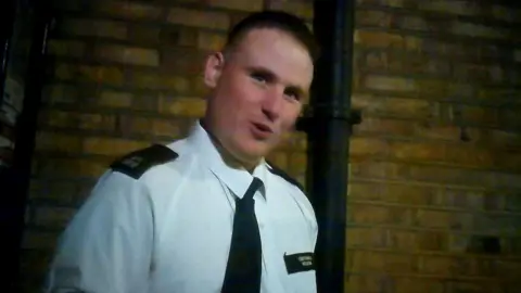 BBC PC Philip Neilson looks at the camera while wearing police uniform comprising of white shirt and tie with shoulder patches in a room against a brick wall, he's slightly blurred as the still is a grab from footage.