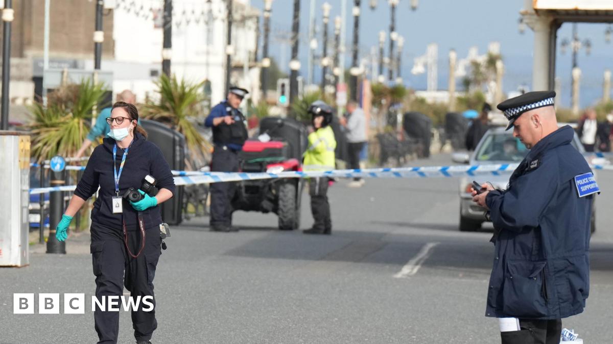 A number of police officers, including a forensics officer holding a camera, on Worthing seafront following a stabbing in the area. A cordon can be seen in the background of the image.