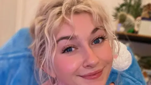 @Eldeeable A woman with blonde hair and blue eyes smiles while looking at the camera. She's in front of a blue fluffy cushion.