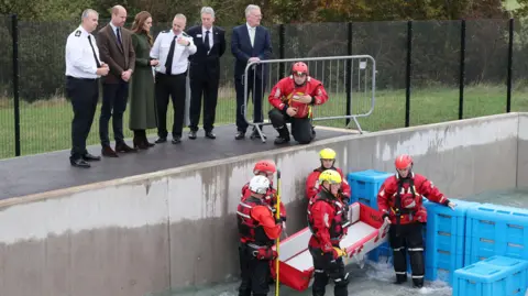 Chris Jackson/PA Wire The Prince and Princess of Wales observe a training scenario during a visit to the Northern Ireland Fire and Rescue Service Learning and Development College near Cookstown. They are standing on a platform with a group of men above a deep training pool.  Five firefighters in red and black rescue uniforms are standing in the pool holding a stretcher as the level of water increases.  