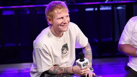 Getty Images Ed Sheeran attends an event in New York on 24 September 2025. He is wearing camouflage trousers and a white T-shirt and has tattoos on his arm. He is holding a microphone and sitting on a box and talking. He has a bottle of water next to him.
