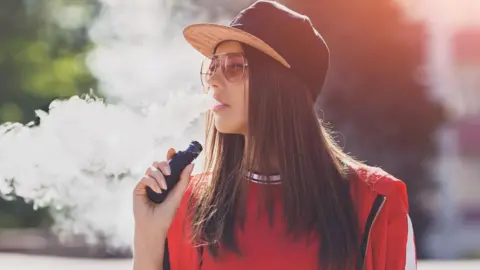 Getty Images A young woman, wearing a cap and sunglasses and dressed in a red top, exhales steam from a vape. She is standing outdoors in the sunshine. 