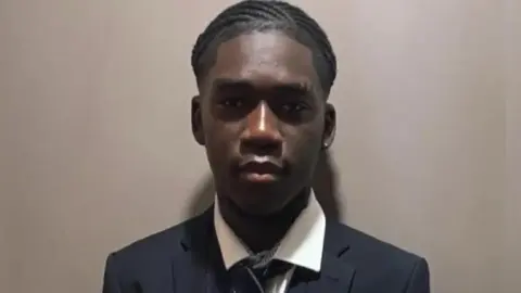 Fakana family Marcus wearing school uniform blazer, shirt and tie. He is standing against a mushroom-coloured wall for a photograph.