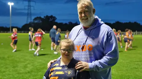 BBC Elsie-May is wearing a Worcester shirt and smiling.  She's standing with her dad, David, on a training pitch.  Women are playing rugby in the background. 