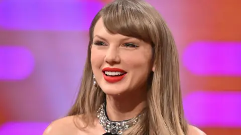 BBC Taylor Swift on the Graham Norton Show, wearing a black dress with a jewelled neckband and smiling, against a purple backdrop