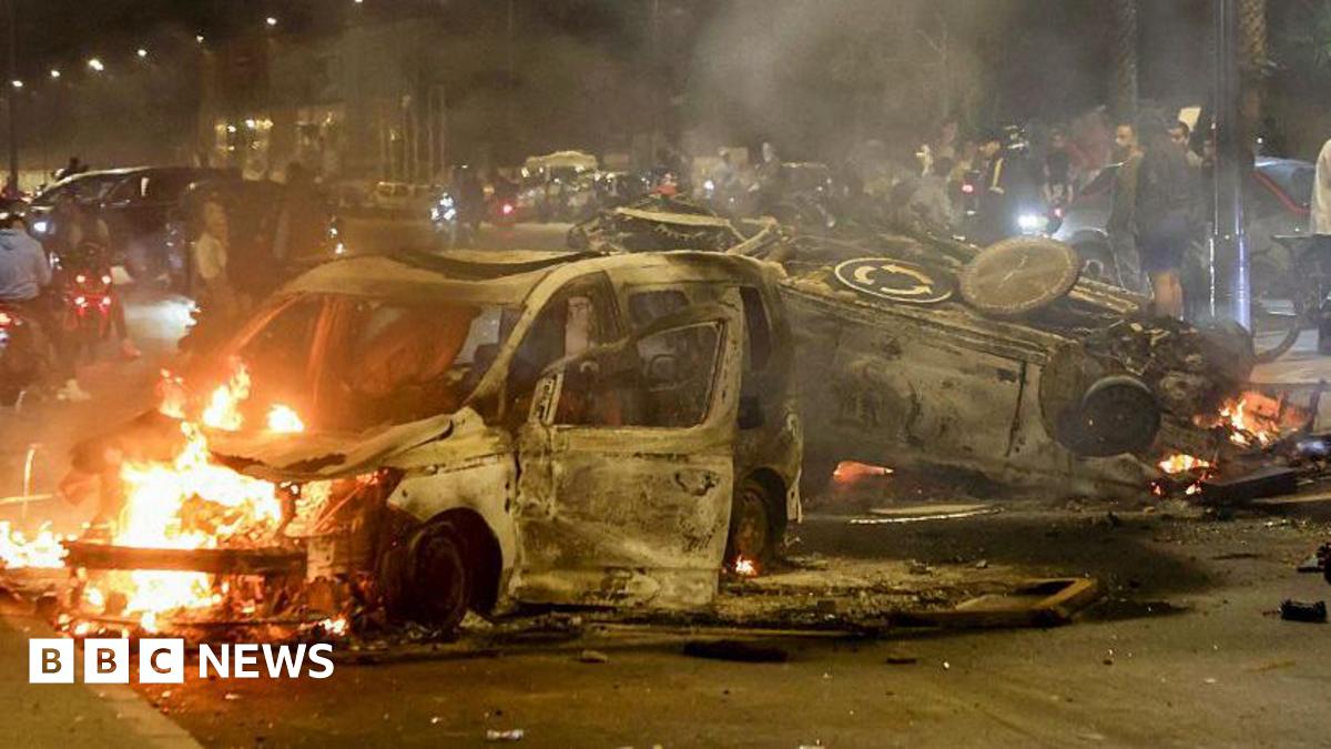 Women walk past police vehicles set on fire during a youth-led demonstration demanding reforms in the healthcare and education sectors in Sale city on 1 October 2025