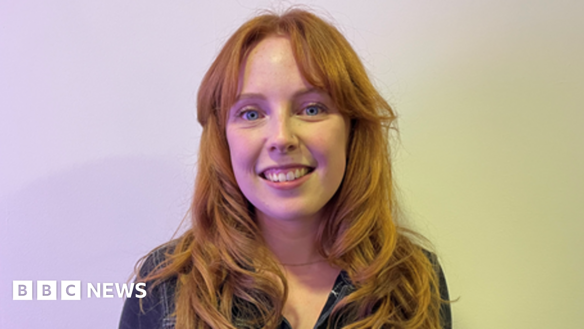 A woman with long red hair, wearing a navy blue jumpsuit, stands in front of a white background. She is smiling into the camera.