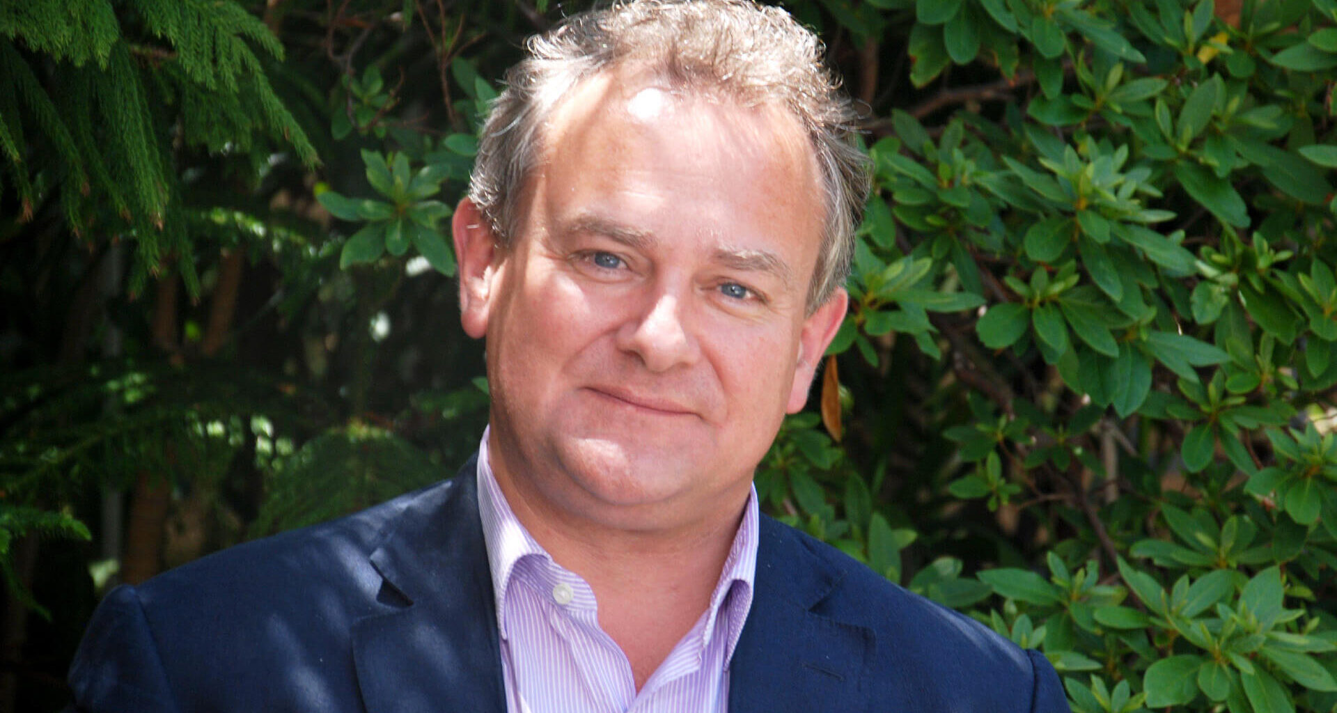 British actor Hugh Bonneville in a photo taken outdoors in front of greenery.