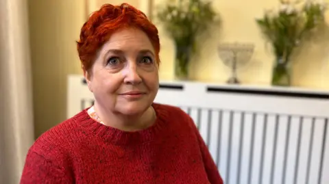 BBC Jennifer Clements wearing a bright red knitted jumper that matches her hair, which is short. Behind her there are vases and flowers and a Hanukkiah candlestick. 
