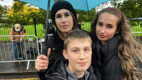 A woman and her two children stand looking into the camera. The woman has drak hair and is wearing a black woolly hat and a black coat. Her daughter has long brown hair and is wearing a black coat. Her son has short brown hair and is wearing a kippah and a black coat. The woman is holding a slight blue umbrella over them, and there are railings and a park behind them.