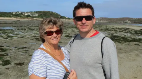 Mark Buzza A man and woman posing for a photo on a beach with blue skies behind.