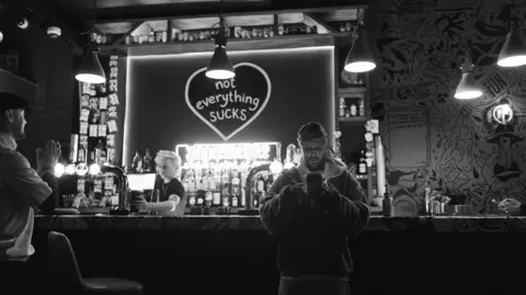 Justin Bieber/Instagram Justin Bieber leaning against a bar, looking at his phone. A graphic behind the bar says "not everything sucks" inside a heart. A barman is serving a pint to another customer. The image is in black and white. 
