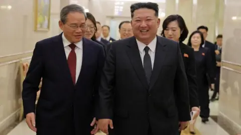 Reuters China's Premier Li Qiang (left) walks beside a smiling Kim Jong Un down a white marble-clad hallway inside a building in Pyongyang. An entourage of officials follows behind them. Everyone is dressed in black suits.