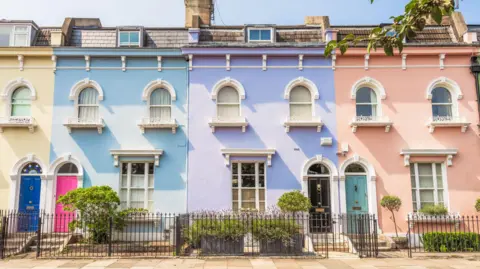 Getty Images A row of terraced houses painted, left to right, yellow, blue, purple and pink, with fences and shrubs in front.