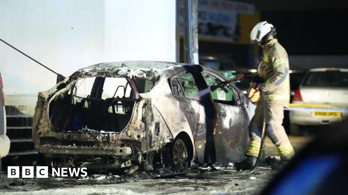 A car is scorched as a firefighter sprays it with water.