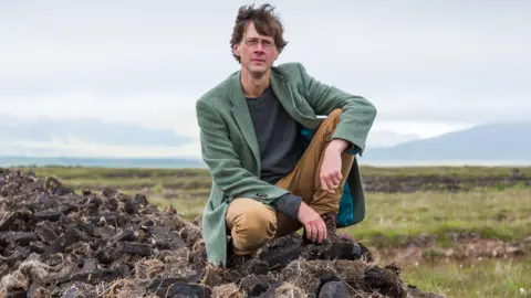 RTE A man in a long green jacket, brown trousers and a blue jumper. He has brown hair. There are mountains behind him. He is kneeling down.
