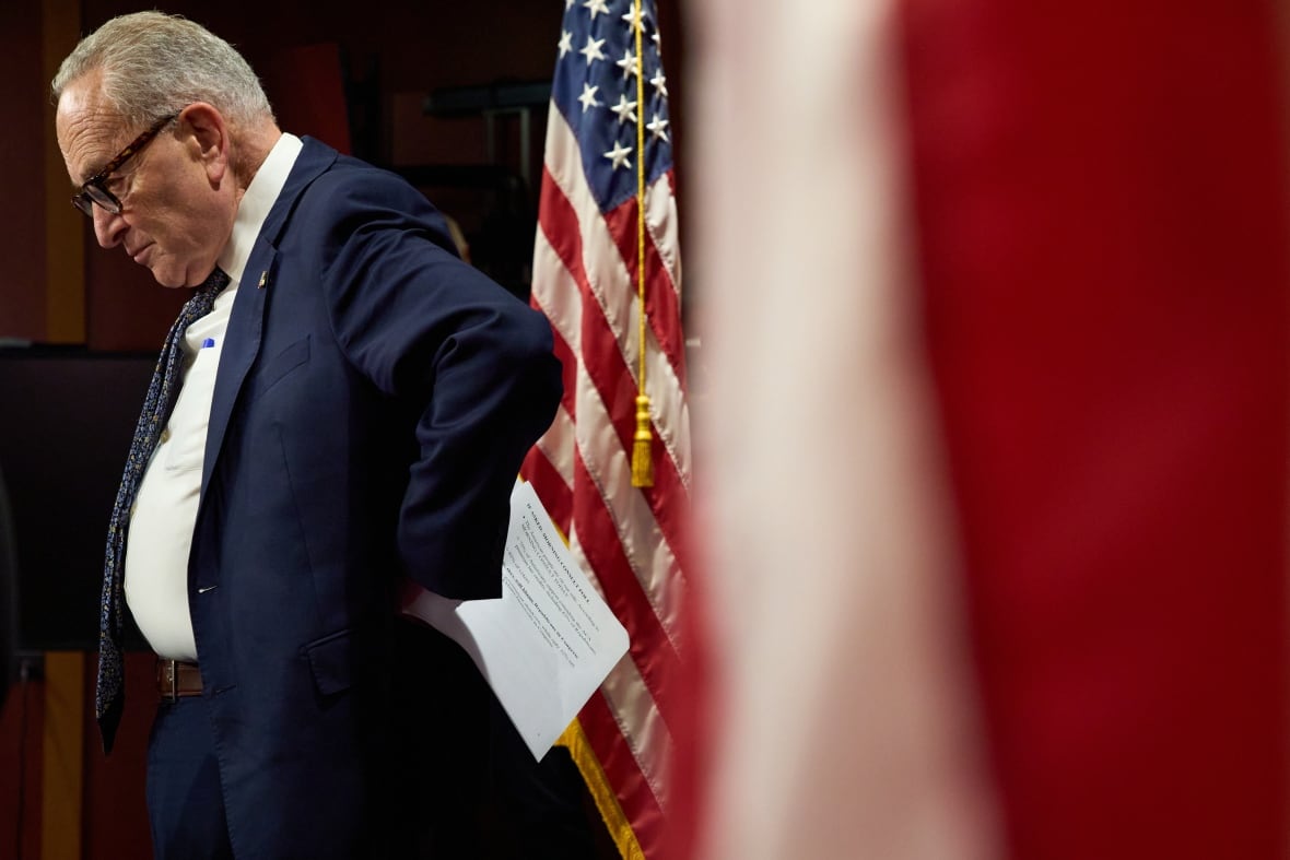 A profile view of a man in a blue suit looking downward with a sullen face as he stands with his hand on his lower back, holding papers, with a red, white and blue U.S. flag behind him. 