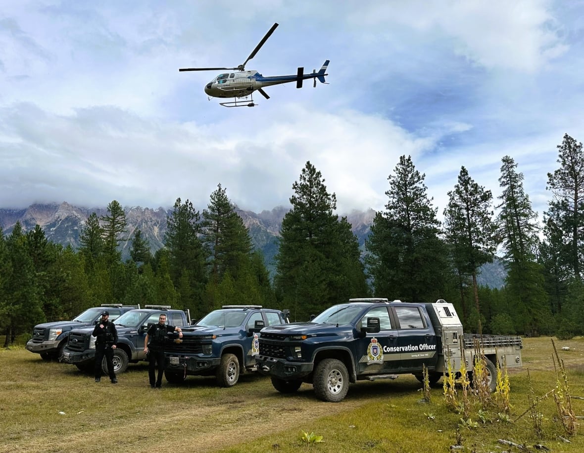 A chopper flies overhead as four patrol cars are parked in a grassland with pine trees and mountains in the background.