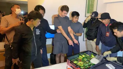 ViralPress A man in a grey shirt and grey shorts. He is looking down and pointing at a suitcase on a bed containing packages in green foil. He is handcuffed. He is surrounded by seven other men - they appear to be Thai police officers in plain clothes. 