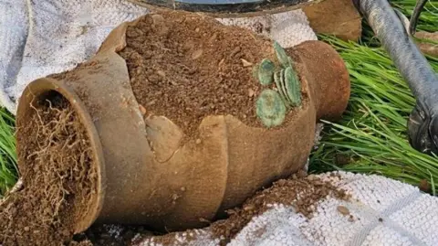 Minelab A clay pot with discoloured coins coming out of it.