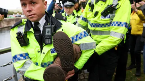 Reuters A group of policemen carry a person by the legs as they're dragged from a Palestinian protest in Liverpool. A huddle of cameramen shoot pictures on either side of the policemen. 