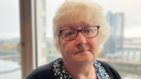 BBC Laura Schosloff is staring at the camera. She has grey hair and glasses and is wearing a patterned black and white blouse and a black cardigan