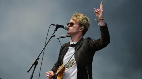 BBC Singer Steve Garrigan wears a brown jacket over a white t-shirt and a pair of sunglasses. He is singing into a microphone and playing a guitar. 
