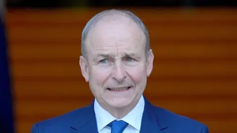 PA Media Micheál Martin stands with a neutral expression on his face. The background is slightly blurred. He is an older man with grey hair and is wearing a navy blue suit with blue tie and white shirt.