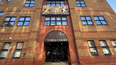 Julita Waleskiewicz/BBC St Albans Crown Court, view of the brown and orange brick building from the outside, with ''Crown Court'' written above the rotating black doors. 