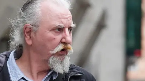 PA Media Paul Martin, a 60-year-old man with long grey hair tied back and a grey and ginger scruffy beard, is looking from the left of the image to the right. He wears a ticking-style shirt, open at the neck, and a black coat.