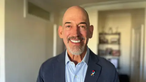 George Carden/BBC Roly Woods wearing a pale blue and white checked shirt and navy blazer, he has a special badge from the Navy which has a Pride flag on it. He is bald and has a beard. He is smiling at the camera. The background is blurred, but appears to be his home, with a shelving unit covered in items visible but blurred behind him