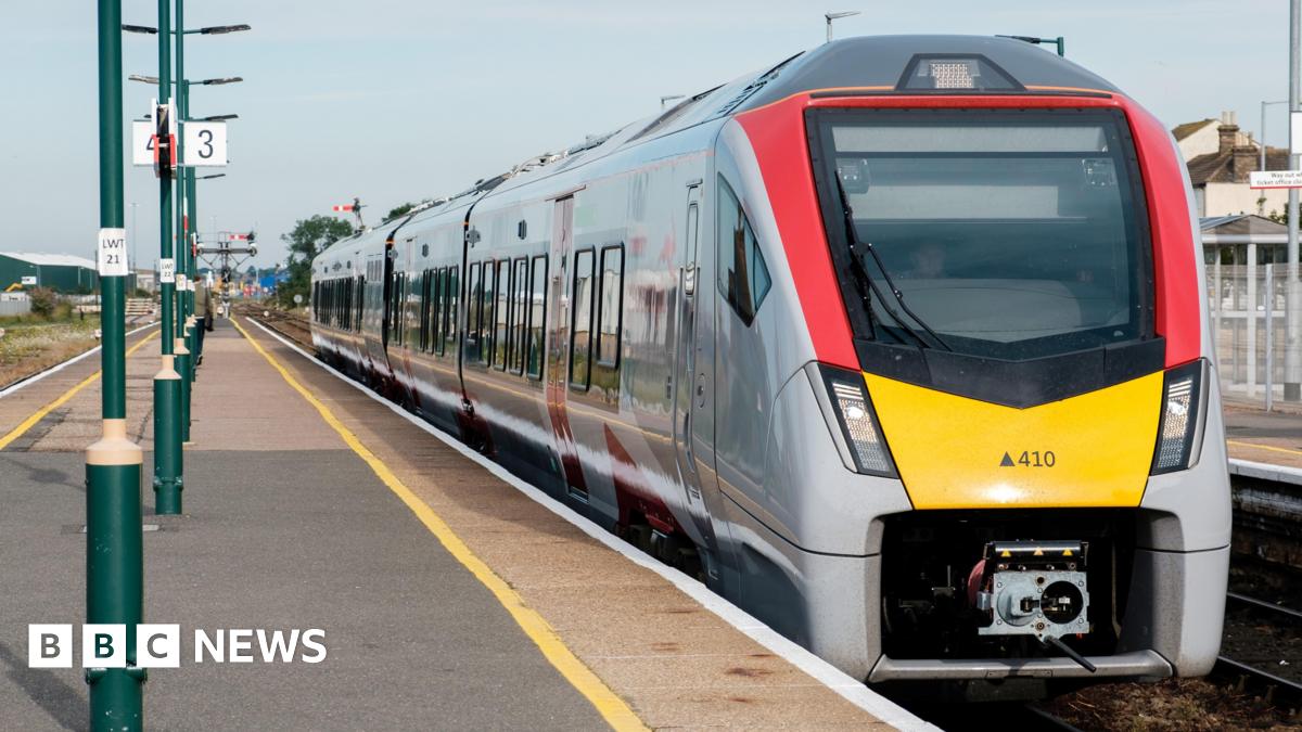 A Greater Anglia train drawing into a station. The platform is in the open and has a number of lamp posts running up the middle of it.