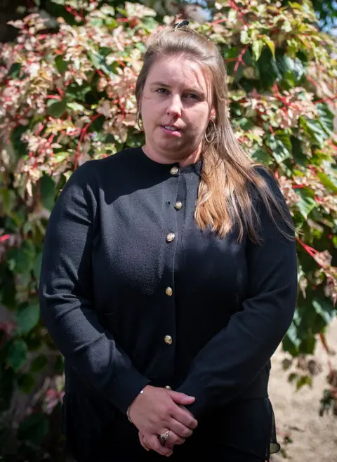 Photo of April Palmer, sister of Alice Webb who died following a Brazilian butt lift procedure one year ago. April is wearing all black clothes and is standing in the sunshine in a garden