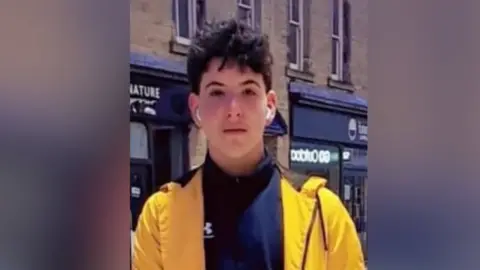 Family handout A teenage boy wearing wireless earphones, a yellow coat and a black tracksuit. He is standing in a town street.