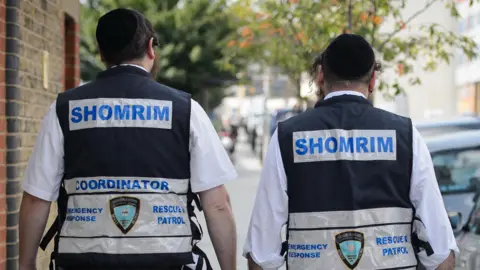 Leon Neal/AFP The back of two Jewish men wearing vests labelled Shomrim across the back walk through north London in 2014. They are wearing white shirts underneath the vests and kippahs.