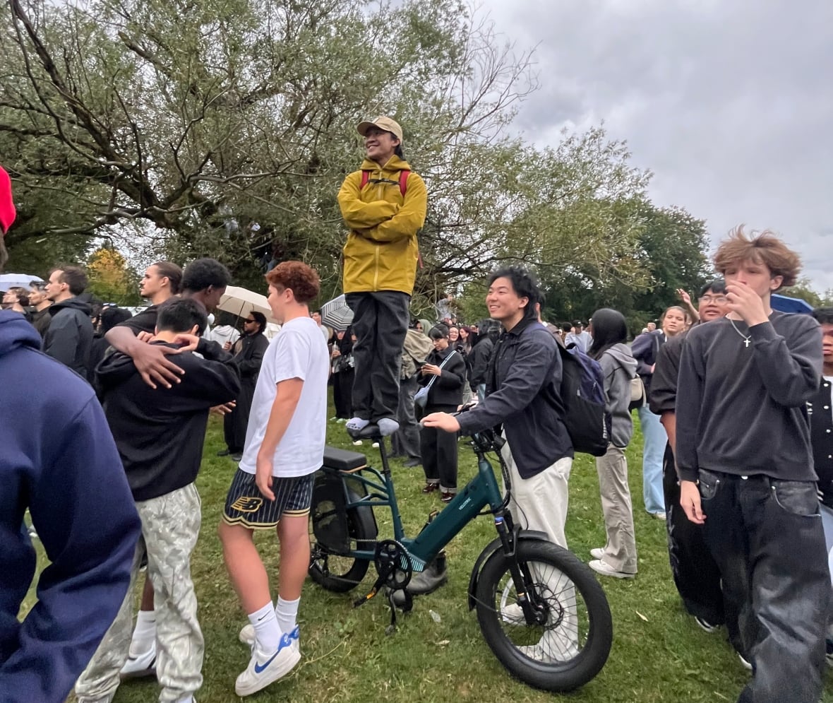 A man stands on his bicycle seat as his friend holds the bike steady, amid a crowd in a park.