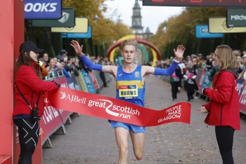 Great Scottish Run Jamie Burns in blue running gear breaks the ribbon as he wins the men's half marathon