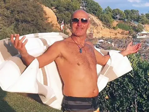 A bald man wearing aviator sunglasses. He has no shirt on and both his arms are splayed out, his lower arms attached to what looks like a white shirt or towel. The sun is shining brightly.