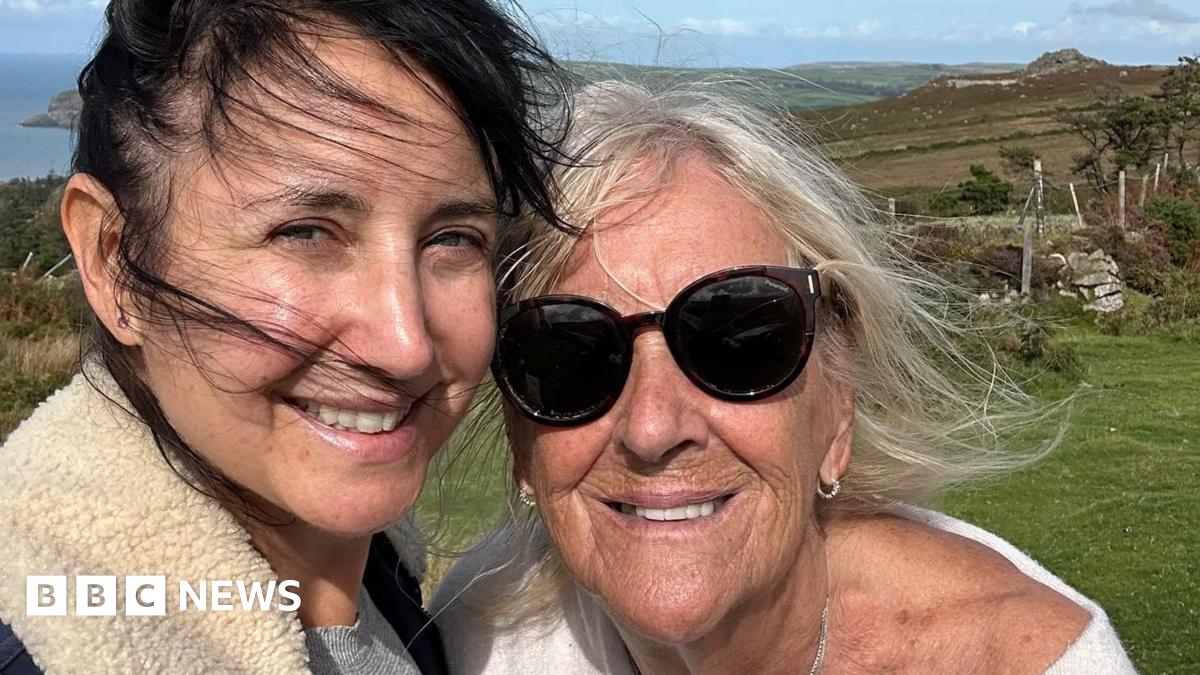 Lucy Owen and her mum Patsy smile in a selfie taken on a coast path in Wales. In the close up photo, Lucy is on the left and is wearing a jacket with a sheepskin collar over a grey jumper. Her dark brown hair is tied up and is blowing across her face as she smiles into the camera. Her mum Patsy Cohen is on the right and is wearing a white jumper with a dropped shoulder, diamond earrings and dark sunglasses. Her white hair is blowing in the wind and the sea is visible behind them.