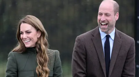 Chris Jackson/PA Wire Catherine, Princess of Wales and her husband Prince William smile and laugh during a rope training exercise at the Northern Ireland Fire and Rescue Service (NIFRS) Learning and Development College in County Tyrone.   Cathering is wearing a dark olive green coat fastened to the neck and Prince William is wearing a brown blazer, a light coloured shirt and a dark tie. 