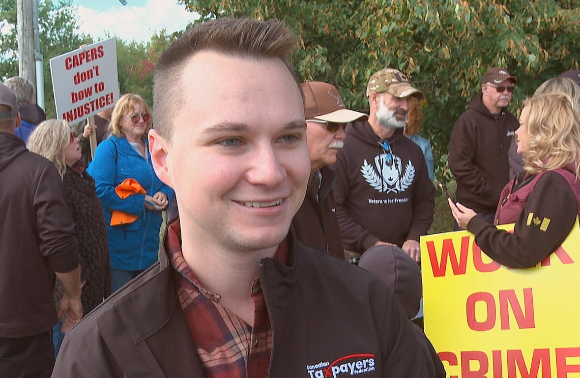 A young man with short spiky hair wearing a black jacket stands in front of some people carrying signs.
