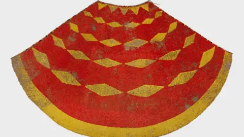 Royal Collection Enterprises Ltd A 19th century yellow and red cloak made from feathers. It has an simple diamond shaped pattern and a yellow border. The feathers have fallen away in parts of the cloak.