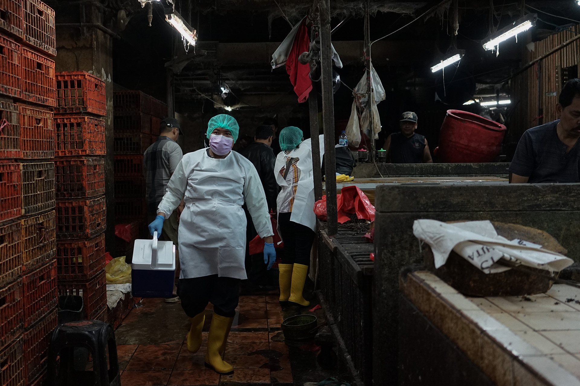 Person in protective gear carrying a cooler in a dimly lit market with stacked crates and hanging lights.