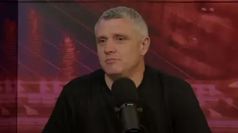 Sunday World Crime World Podcast/YouTube A screenshot of Niall Donald sitting in front of a black, table microphone while recording the Crime World podcast. Mr Donald is an older man with short, greying hair and is wearing a black t-shirt. He is sitting in front of red screen, displaying the Samuel Beckett bridge.